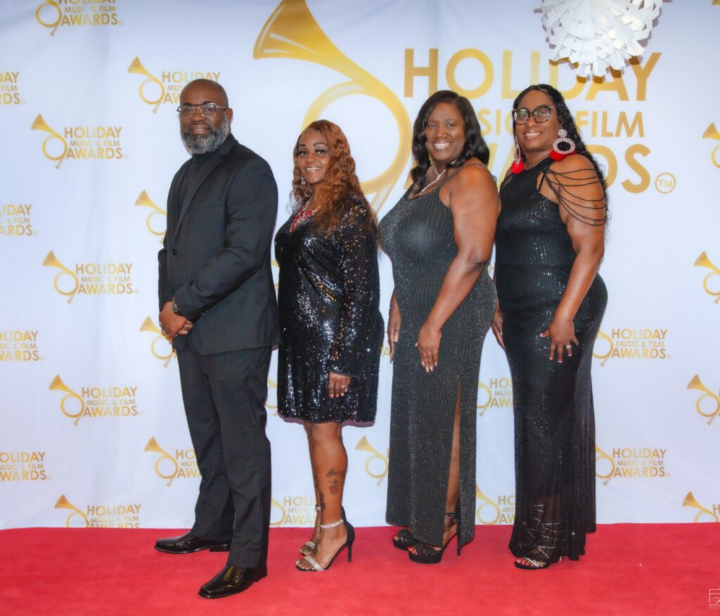 Four people standing on a red carpet in front of a white wall.